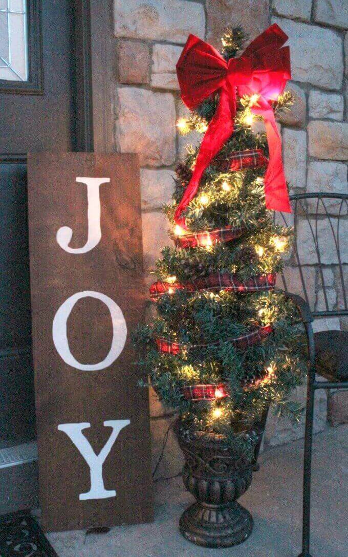"Joy" painted on rustic wood sign with small outdoor lighted Christmas tree on front porch.