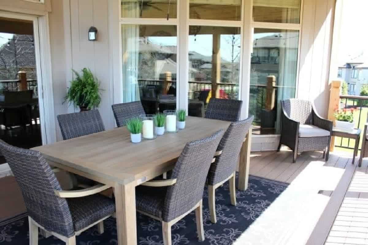 Composite deck with black and tan outdoor rug, teak table, and wicker chairs.