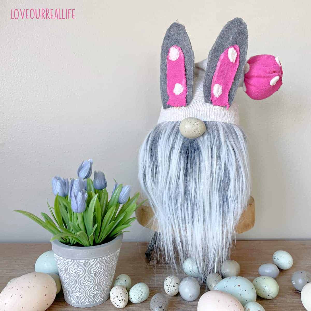 Easter bunny gnome sitting on stand beside faux tulips with plastic eggs on ground.