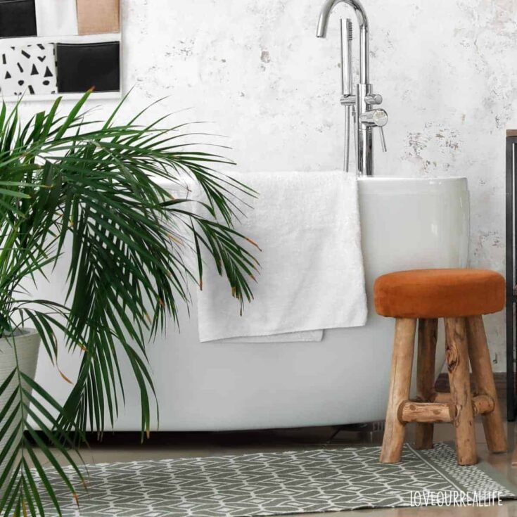 Green and white cotton blend bathroom rug beside a large white tub with plant and stool.