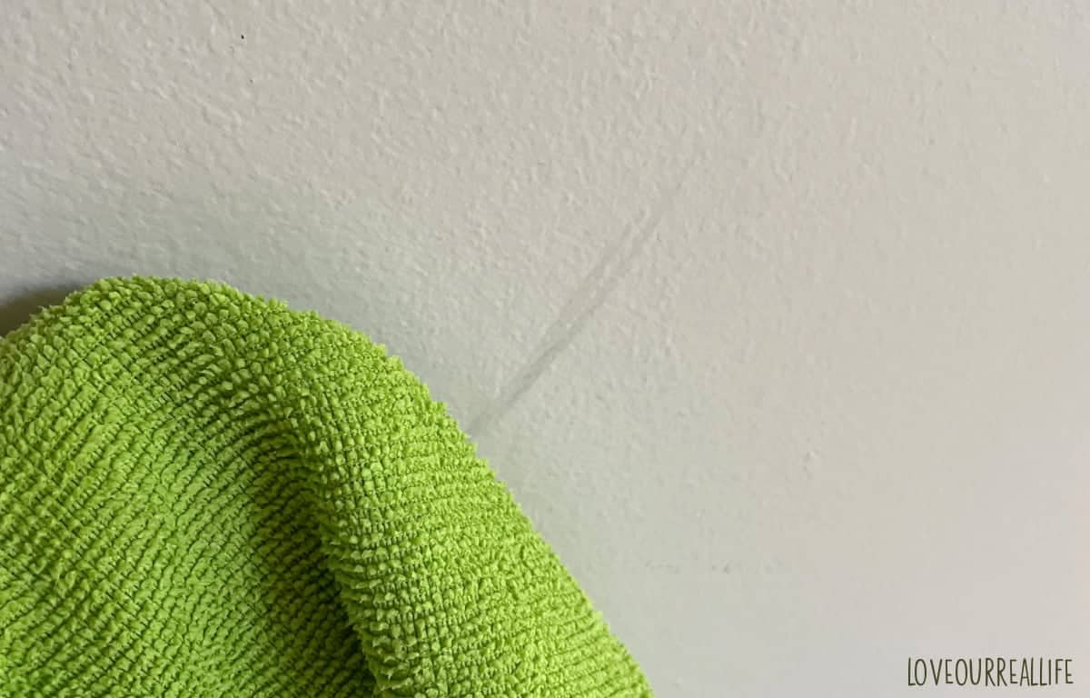 Cleaning scuff mark on gray painted wall using green microfiber cloth.