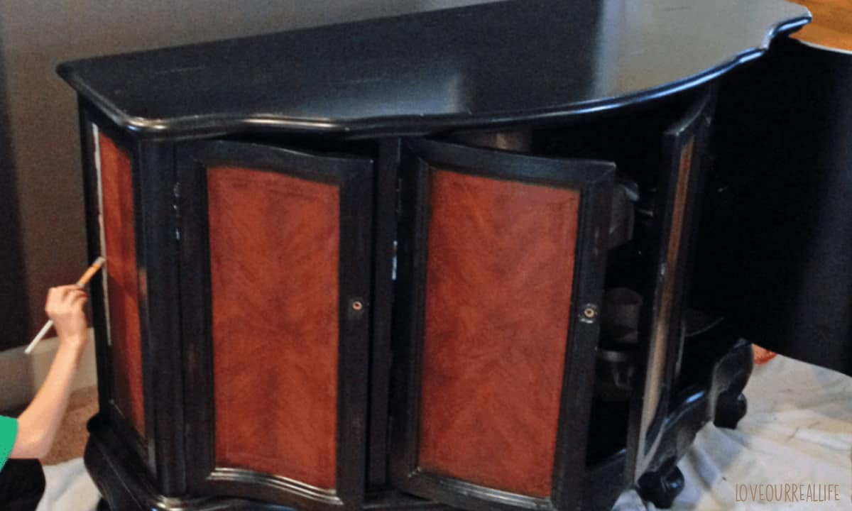 Black and brown cabinet with doors open, with someone holding a paintbrush to paint.
