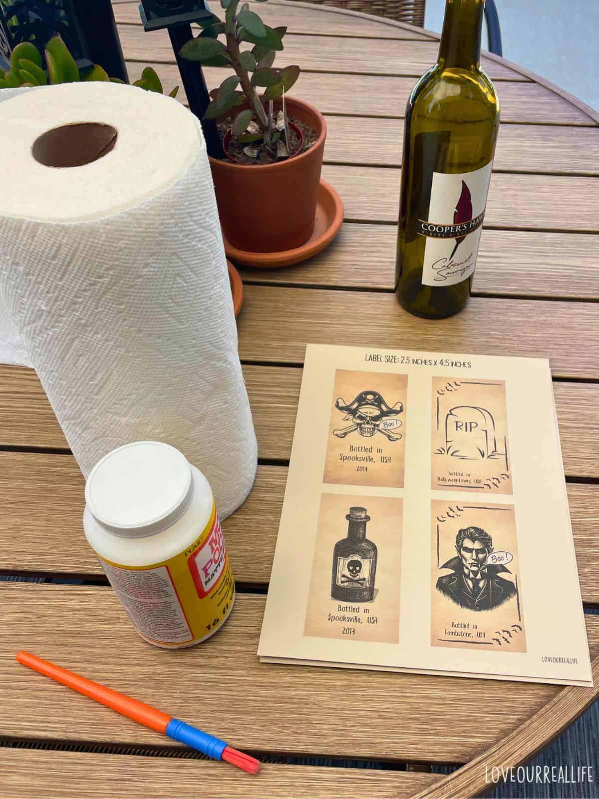 Paper towels, Mod Podge, wine bottle, and halloween bottle labels sitting on outdoor table.
